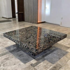 Puede incluir: Una mesa de centro cuadrada con una superficie oscura y estampada. La mesa tiene un perfil bajo y una base gruesa y sólida. La superficie parece estar hecha de piedra pulida con un patrón único y orgánico en tonos de negro, gris y crema.