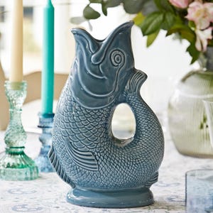 May include: A blue ceramic fish-shaped jug with scale and fin details. The jug has a handle and a wide opening. The background features candles, flowers, and a patterned tablecloth. The jug is approximately 25 cm tall.