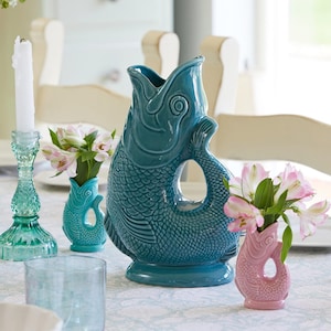 May include: A teal ceramic fish-shaped pitcher with a handle and a wide opening, alongside smaller fish-shaped vases in teal and pink, each holding pink and white flowers. A teal glass candlestick and a clear glass tumbler are also present.