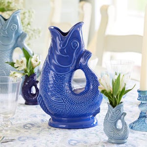 Könnte beinhalten: Eine Sammlung blauer Keramik-Fischkrüge und Vasen. Der größte Krug ist tiefblau, mit einem Griff und einem Design mit offenem Mund. Kleinere Vasen in verschiedenen Blautönen halten weiße Blumen. Die Fischschuppenstruktur ist am Krugkörper sichtbar.