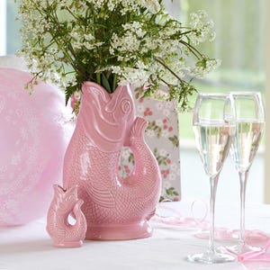 Könnte beinhalten: Eine rosa Keramikvase in Fischform mit weißen Blumen. Eine kleinere passende Vase steht davor. Zwei Sektgläser mit einem prickelnden Getränk befinden sich rechts. Rosa Bänder und ein rosa Kissen im Hintergrund.