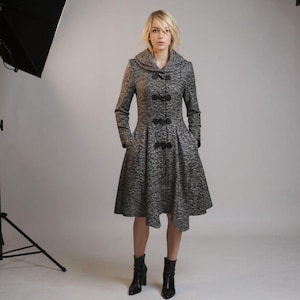 May include: A gray textured coat with a flared skirt and a high collar. The coat features black buttons and pockets. The model is wearing black ankle boots.