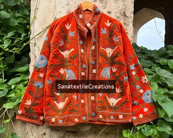 May include: An orange velvet jacket with intricate floral embroidery in white, blue, and green. The jacket features a mandarin collar and is displayed on a wooden hanger. The text "SanatextileCreations" is visible.