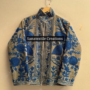 Op de afbeelding: Een beige jas met levendige blauwe bloemenborduursels op de voorzijde en de mouwen. Het kledingstuk heeft een hoge kraag en lange mouwen. De tekst Sanatextile Creations is in het zwart in het midden op de voorzijde van de jas gedrukt.