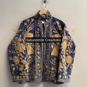 Pode incluir: Uma jaqueta azul com intrincados bordados florais dourados nos painéis frontais e nas mangas. A peça tem gola alta, mangas compridas e um design aberto na frente. O texto Sanatextile Creations é visível no centro.