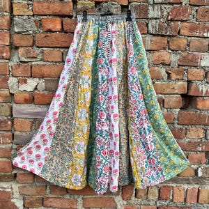 May include: A long, flowing skirt with a patchwork design. The skirt features various floral and geometric patterns in shades of white, pink, yellow, green, and brown. It is hanging against a weathered brick wall.