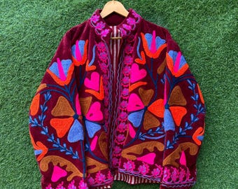 May include: A burgundy velvet jacket with vibrant floral embroidery in shades of pink, orange, blue, and brown. The jacket features a high collar and a striped lining, creating a striking contrast. The embroidery is detailed and covers the entire jacket.