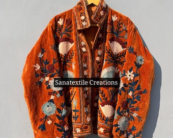 May include: An orange velvet jacket featuring intricate floral embroidery in shades of blue, white, and cream. The garment has long sleeves, a high collar, and a front opening. The text Sanatextile Creations is printed across the center front.