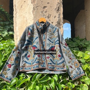Pode incluir: Uma jaqueta cinza com bordados florais intrincados em vermelho, azul e dourado. A jaqueta apresenta gola alta e mangas compridas, com o texto "SanatextileCreations" visível. A jaqueta está pendurada em um cabide de madeira.