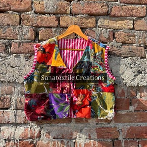 Op de afbeelding: Een kleurrijk patchwork vest met een V-hals en een trekkoordsluiting. Het vest is gemaakt van verschillende stoffen vierkanten. De binnenvoering is roze en wit gestreept. De tekst "Sanatextile Creations" is op de voorkant gedrukt.