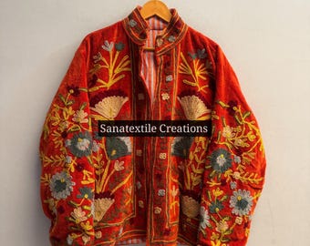 May include: A vibrant orange velvet jacket featuring intricate floral embroidery in shades of yellow, grey, and cream. The garment has a high collar, long sleeves, and a front opening. The text Sanatextile Creations is printed across the center.