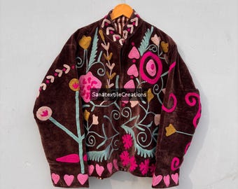 以下が含まれることがあります： 茶色のベルベットジャケット、カラフルな花の刺繍入り。 ピンク、黄色、ターコイズブルーの刺繍の花と葉が特徴です。 ジャケットは、明るい灰色の壁を背景に、木製のハンガーに掛けられています。 「SanatextileCreations」の文字が見えます。