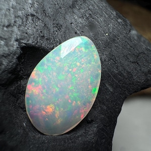 May include: A pear-shaped opal gemstone featuring a translucent white base with vibrant flashes of red, orange, and green color play, resting against a dark, textured, charcoal-like surface that highlights the stone's internal brilliance.