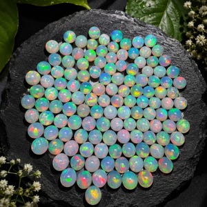 Op de afbeelding: Een verzameling van talloze kleine, ronde, doorschijnende cabochon-opalen. Elke edelsteen vertoont een melkachtige witte basiskleur met levendige, iriserende flitsen van groen, oranje en blauw licht over hun gladde, gepolijste oppervlakken.
