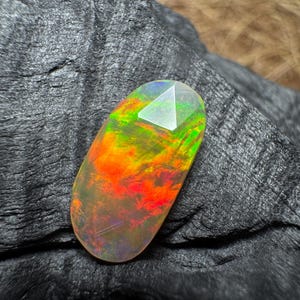 May include: An oval-shaped faceted gemstone displaying a vibrant play of color with intense flashes of neon orange, bright red, and electric green against a translucent base, showcasing a polished surface with geometric facets.