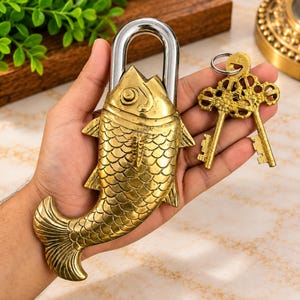 Könnte beinhalten: Ein fischförmiges Vorhängeschloss aus Messing mit einem silberfarbenen Metallbügel und zwei passenden, verzierten Messingschlüsseln an einem Ring. Das Schloss zeigt detaillierte Schuppenstrukturen, ein Auge sowie eine geschwungene Schwanzflosse.