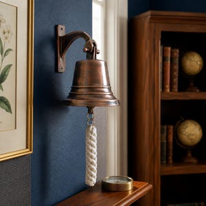 May include: A bronze-colored bell with a rope pull hangs on a dark blue wall. The bell is mounted on a curved bracket. A white rope pull hangs from the bell. The bell is part of a nautical or vintage decor theme.