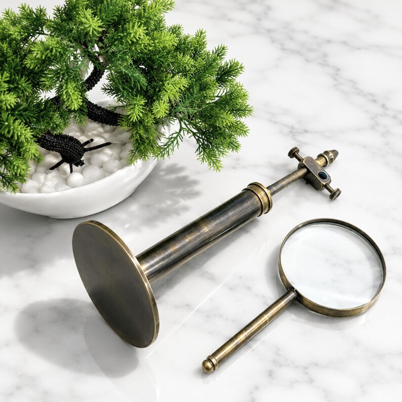 Pu&ograve; includere: Un telescopio e una lente d'ingrandimento in ottone antico su una superficie di marmo. Un piccolo bonsai decorativo in una ciotola bianca &egrave; sullo sfondo. Il telescopio ha una base grande e rotonda e una messa a fuoco regolabile. La lente d'ingrandimento ha una lente rotonda.