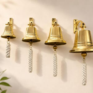 Pode incluir: Um conjunto de quatro sinos de lat&atilde;o polido de tamanhos variados, cada um montado num suporte de parede. Cada sino apresenta uma superf&iacute;cie dourada lisa e reflexiva, equipada com uma corda tran&ccedil;ada branca presa ao badalo central.
