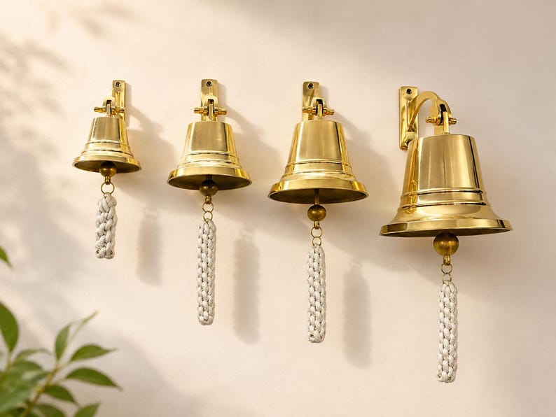 Pode incluir: Um conjunto de quatro sinos de lat&atilde;o polido de tamanhos variados, cada um montado num suporte de parede. Cada sino apresenta uma superf&iacute;cie dourada lisa e reflexiva, equipada com uma corda tran&ccedil;ada branca presa ao badalo central.