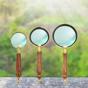 Può includere: Tre lenti d'ingrandimento con montature dorate e manici in legno. Le lenti sono chiare e i manici hanno un design intagliato dettagliato. Lo sfondo è sfocato in verde e bianco, suggerendo un ambiente esterno. Le lenti d'ingrandimento sono di dimensioni variabili.