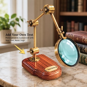 Può includere: Una lente d'ingrandimento color ottone con base in legno. Il testo "Add Your Own Text" e "Text here" è visibile. La lente d'ingrandimento è regolabile e ha una lente trasparente. La base è in legno scuro.