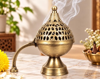 May include: A brass-toned metal incense burner with a dome-shaped lid featuring intricate cut-out patterns. The burner has a curved handle, a pedestal base, and emits a visible trail of white smoke from the top finial.