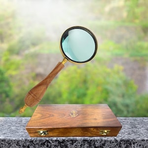 May include: A magnifying glass with a wooden handle and brass accents is positioned above a wooden box. The box has a brass clasp and an anchor emblem. The background is a blurred view of greenery.