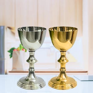 Puede incluir: Se muestran dos cálices ornamentados, uno plateado y otro dorado. Los cálices tienen una forma de cuenco redondeada, un tallo detallado y una base circular. El cáliz plateado está a la izquierda y el dorado a la derecha.