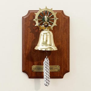 Könnte beinhalten: Eine Schiffsglocke aus Messing, montiert auf einer dunklen Holzplatte. Die Glocke ist mit einem dekorativen Steuerrad versehen und hat einen geflochtenen Seilzug. Eine Messingplatte darunter trägt die Aufschrift "S.S. ATLANTA EST. 1912".