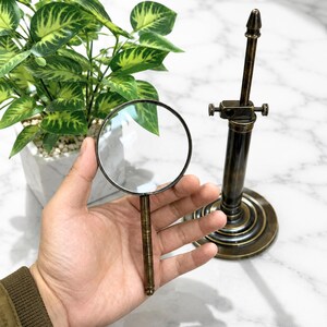 Pu&ograve; includere: Una lente d'ingrandimento in stile vintage con manico e montatura in ottone, tenuta in mano. Accanto, un supporto in ottone abbinato con elementi regolabili. Lo sfondo include una pianta in vaso e una superficie marmorizzata.