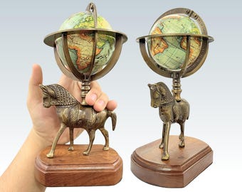 May include: Two antique-style globes with a bronze finish, each mounted on a horse figurine base. The globes feature a detailed map design with shades of green and brown. The horse figurines are set on a rectangular wooden base.