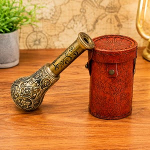 Può includere: Un telescopio in ottone con un motivo floreale decorato sull'impugnatura e un tubo cilindrico con testo inciso, abbinato a una custodia cilindrica in pelle marrone goffrata con motivi nautici, inclusi timoni e disegni di bussole.