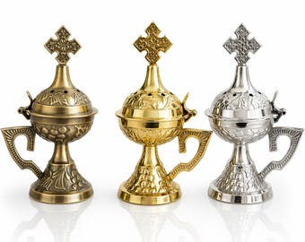 May include: Three ornate metal incense burners in gold, bronze, and silver. Each burner features a cross-shaped top, a round body with decorative patterns, a handle, and a pedestal base. The burners are displayed on a white surface.
