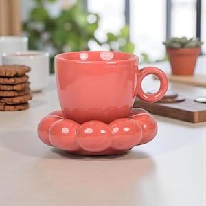 Puede incluir: Una taza de cerámica de color coral con un asa redondeada se encuentra sobre un posavasos a juego con forma de flor. La taza y el posavasos están sobre una superficie blanca, con galletas y otras tazas en el fondo.