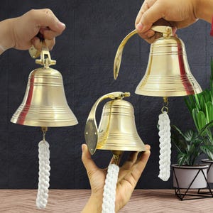 Può includere: Tre campane di nave in ottone con manici curvi e tiranti in corda bianca. Le campane hanno una finitura lucida e sono montate su uno sfondo scuro. Un oggetto decorativo nautico.