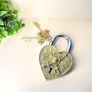 Puede incluir: Un candado en forma de corazón con un diseño ornamentado y grabado en tonos dorados y plateados. Dos llaves decorativas están junto al candado. El grillete del candado es plateado y azul. El fondo es de color beige claro.