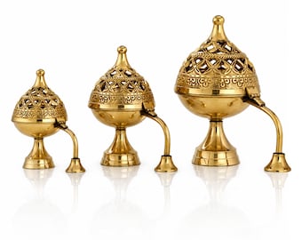 May include: Three brass incense burners of varying sizes. Each burner has a domed, pierced lid, a rounded body, and a pedestal base. A curved handle extends from the body. The burners are gold-colored and sit on a white surface.