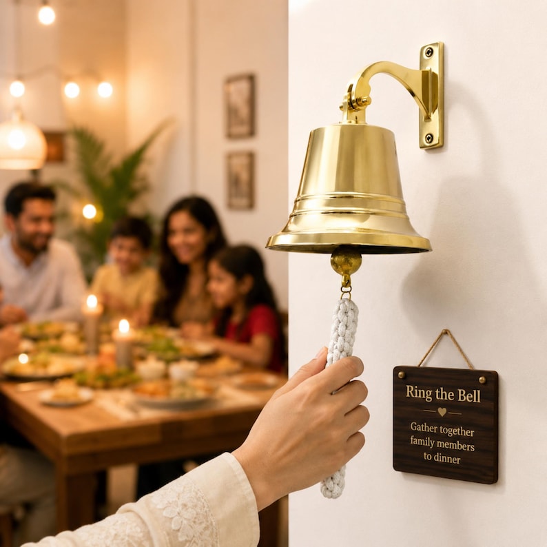 Pode incluir: Um sino de parede em lat&atilde;o polido com um puxador de corda tran&ccedil;ada branca. Abaixo do sino, pendura-se uma placa retangular de madeira escura com o texto "Ring the Bell" e "Gather together family members to dinner" gravado em cor clara.
