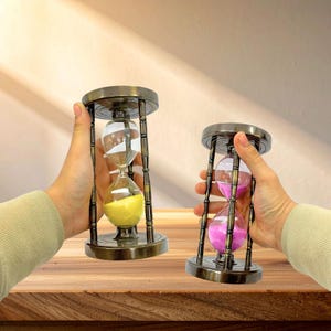 May include: Two hourglasses with bronze-coloured metal frames are held in hands. One contains yellow sand, the other pink sand. The glass bulbs are supported by a decorative metal base and top. The hourglasses are on a wooden surface.