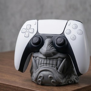 May include: A white and black PlayStation 5 controller sits atop a grey, detailed sculpture of a Japanese demon's face. The controller's buttons and joysticks are visible. The sculpture is on a wooden surface.