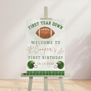 Könnte beinhalten: Ein Schild für einen ersten Geburtstag mit Football-Motiv. Es zeigt den Text "First Year Down", "Welcome to Cooper's First Birthday" und das Datum. Es enthält einen Football, Helme und ein Feld.