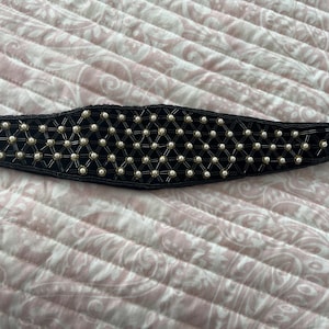 May include: Black beaded belt with a velvet backing. The belt features a geometric pattern of small, round, off-white beads. The belt is a wide style, suitable for waist or hip wear. The belt is on a pink and white patterned surface.