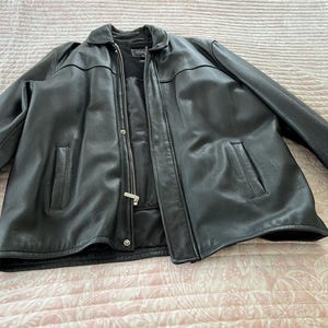 Puede incluir: Chaqueta de cuero negra con cuello, dos bolsillos delanteros y cierre de cremallera. La chaqueta está tendida sobre una superficie estampada. La chaqueta tiene un diseño clásico.