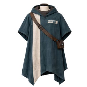 Puede incluir: Poncho color turquesa con una franja vertical crema, capucha y bandolera de cuero marrón. El poncho tiene la palabra "ARE" en un parche rectangular. La prenda se exhibe en un maniquí.