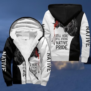 Könnte beinhalten: Ein schwarz-weißer Kapuzenpullover mit Reißverschluss, der ein Wolfsmotiv und den Text "STILL HERE STILL STRONG NATIVE PRIDE" zeigt. Das Wort "NATIVE" ist vertikal auf den Ärmeln aufgedruckt. Der Hoodie hat ein weißes Innenfutter.