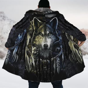 Könnte beinhalten: Ein schwarzer Kapuzenmantel mit einem Wolfsmotiv. Das Kunstwerk zeigt ein großes Wolfsgesicht mit stechenden Augen, umgeben von Federn und anderen Wolfsbildern. Der Umhang hat einen dunklen, strukturierten Hintergrund und ein weiches, graues Innenfutter.