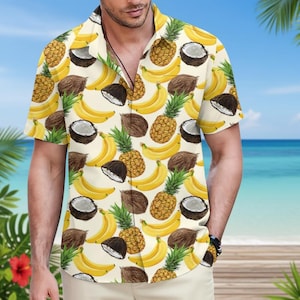 Könnte beinhalten: Kurzärmeliges Hemd mit tropischem Fruchtmuster. Das Hemd zeigt ein sich wiederholendes Design aus gelben Bananen, Ananas und Kokosnüssen auf cremefarbenem Hintergrund. Das Hemd wird von einer Person getragen.