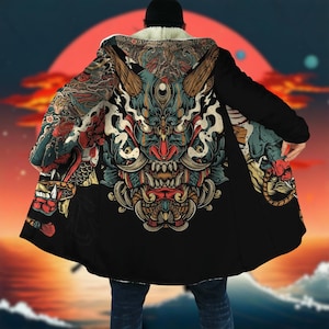 Puede incluir: Abrigo negro con capucha que presenta una detallada y colorida ilustración de una máscara de demonio japonés en la espalda. El abrigo tiene un forro blanco y esponjoso alrededor de la capucha y un fondo de puesta de sol.