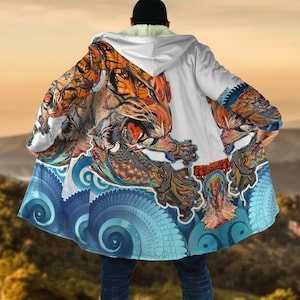 Könnte beinhalten: Weißer Kapuzenmantel mit einem lebendigen Tigerdesign. Der Tiger ist mit orangefarbenen, braunen und blauen Mustern dargestellt. Der Umhang hat unten ein blau-weißes Wellenmuster und ein weiches, weißes Innenfutter.